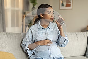 Young african american pregnant lady with big belly drinking glass of water, enjoying free time, sitting on sofa at home