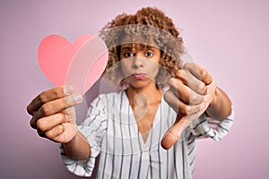 Young african american romantic woman with curly hair holding paper heart shape with angry face, negative sign showing dislike