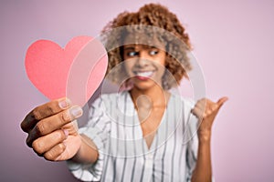 Young african american romantic woman with curly hair holding paper heart shape pointing and showing with thumb up to the side