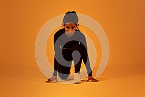 Young african american runner woman doing crouch start running on orange neon studio background, wearing black fitwear