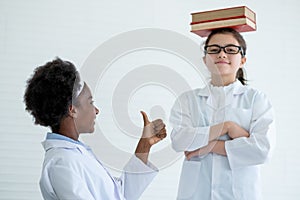 Young African American scientist girl shows thumbs up to her Caucasian friend who put stack of books on her head in laboratory or