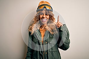 Young african american skier woman with curly hair wearing snow sportswear and ski goggles showing and pointing up with finger