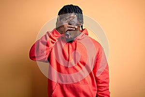 Young african american sporty man wearing sweatshirt with hoodie over yellow background peeking in shock covering face and eyes