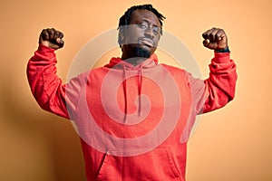 Young african american sporty man wearing sweatshirt with hoodie over yellow background showing arms muscles smiling proud