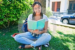 Young african american student woman reading a book sitting on the grass at the university campus