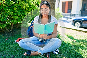 Young african american student woman reading a book sitting on the grass at the university campus