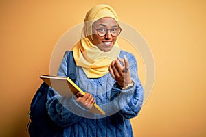 Young African American student woman wearing muslim hijab and backpack holding book Beckoning come here gesture with hand inviting