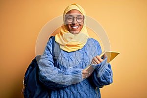 Young African American student woman wearing muslim hijab and backpack holding book with a happy and cool smile on face