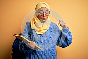 Young African American student woman wearing muslim hijab and backpack holding book Pointing down with fingers showing