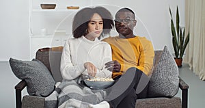 Young african american teen couple black man and ethnic woman family watching movie sitting on sofa at home covered with