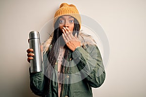 Young african american tourtist woman wearing coat and drinking water from thermo cover mouth with hand shocked with shame for