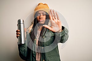 Young african american tourtist woman wearing coat and drinking water from thermo with open hand doing stop sign with serious and