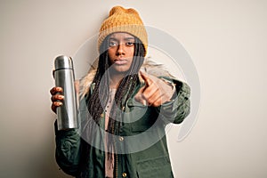 Young african american tourtist woman wearing coat and drinking water from thermo pointing with finger to the camera and to you,