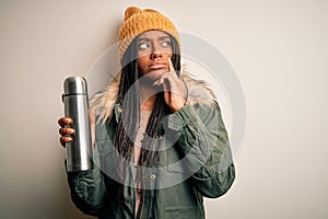 Young african american tourtist woman wearing coat and drinking water from thermo serious face thinking about question, very