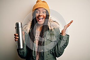 Young african american tourtist woman wearing coat and drinking water from thermo very happy and excited, winner expression