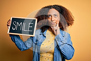 Young african american woman with afro hair holding blackboard with smile message serious face thinking about question, very