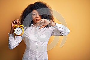 Young african american woman with afro hair holding classic alarm clock over yellow background with angry face, negative sign