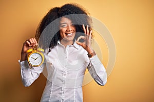 Young african american woman with afro hair holding classic alarm clock over yellow background doing ok sign with fingers,