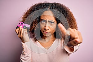 Young african american woman with afro hair holding with hand small pink toy car pointing with finger to the camera and to you,