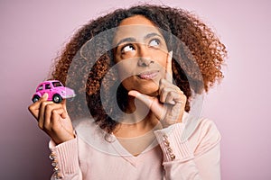 Young african american woman with afro hair holding with hand small pink toy car serious face thinking about question, very