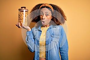 Young african american woman with afro hair holding jar with healthy macaroni pasta scared in shock with a surprise face, afraid