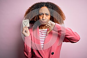 Young african american woman with afro hair holding paper with heart over pink background with angry face, negative sign showing