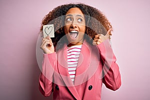 Young african american woman with afro hair holding paper with heart over pink background pointing and showing with thumb up to