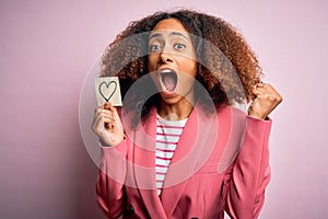 Young african american woman with afro hair holding paper with heart over pink background screaming proud and celebrating victory