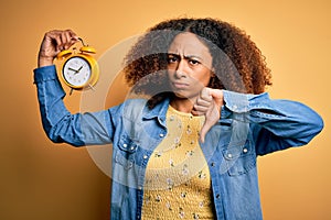 Young african american woman with afro hair holding vintage alarm clock over yellow background with angry face, negative sign