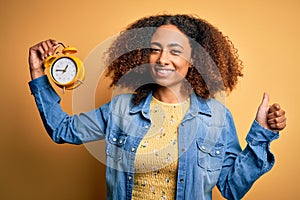 Young african american woman with afro hair holding vintage alarm clock over yellow background happy with big smile doing ok sign,
