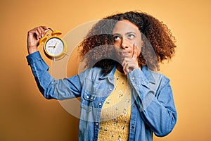 Young african american woman with afro hair holding vintage alarm clock over yellow background serious face thinking about