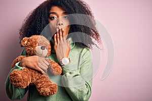 Young african american woman with afro hair hugging teddy bear over pink background cover mouth with hand shocked with shame for