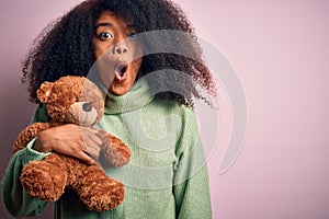 Young african american woman with afro hair hugging teddy bear over pink background scared in shock with a surprise face, afraid