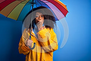 Young african american woman with afro hair under colorful umbrella wearing winter coat for rain cover mouth with hand shocked