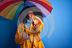 Young african american woman with afro hair under colorful umbrella wearing winter coat for rain pointing with finger to the