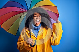 Young african american woman with afro hair under colorful umbrella wearing winter coat for rain screaming proud and celebrating