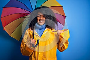 Young african american woman with afro hair under colorful umbrella wearing winter coat for rain with surprise face pointing