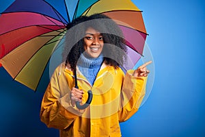 Young african american woman with afro hair under colorful umbrella wearing winter coat for rain very happy pointing with hand and