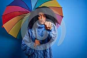 Young african american woman with afro hair under colorful umbrella for winter weather rain pointing with finger to the camera and