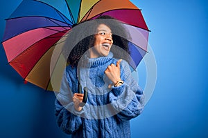 Young african american woman with afro hair under colorful umbrella for winter weather rain pointing and showing with thumb up to