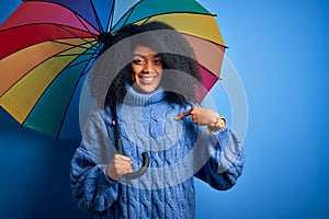 Young african american woman with afro hair under colorful umbrella for winter weather rain with surprise face pointing finger to