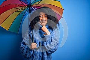Young african american woman with afro hair under colorful umbrella for winter weather rain very happy pointing with hand and