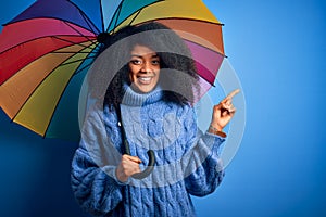 Young african american woman with afro hair under colorful umbrella for winter weather rain very happy pointing with hand and