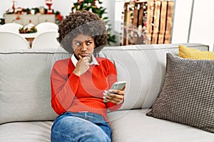 Young african american woman with afro hair using smartphone sitting on the sofa by christmas tree serious face thinking about