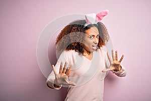 Young african american woman with afro hair wearing bunny ears over pink background afraid and terrified with fear expression stop