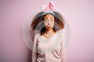 Young african american woman with afro hair wearing bunny ears over pink background puffing cheeks with funny face