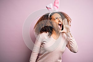 Young african american woman with afro hair wearing bunny ears over pink background shouting and screaming loud to side with hand