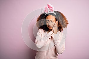 Young african american woman with afro hair wearing bunny ears over pink background sleeping tired dreaming and posing with hands