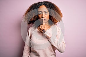 Young african american woman with afro hair wearing casual sweater over pink background asking to be quiet with finger on lips