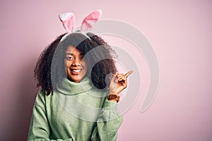 Young african american woman with afro hair wearing easter rabbit ears costume over pink background with a big smile on face,